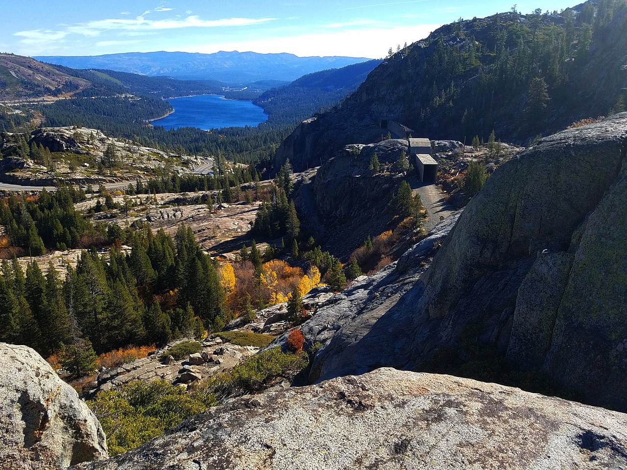 View of Donner Lake surrounded by rugged mountain terrain and lush forests, captured from a high vantage point. The clear blue lake contrasts beautifully with the vibrant autumn foliage and rocky outcrops under a bright blue sky.